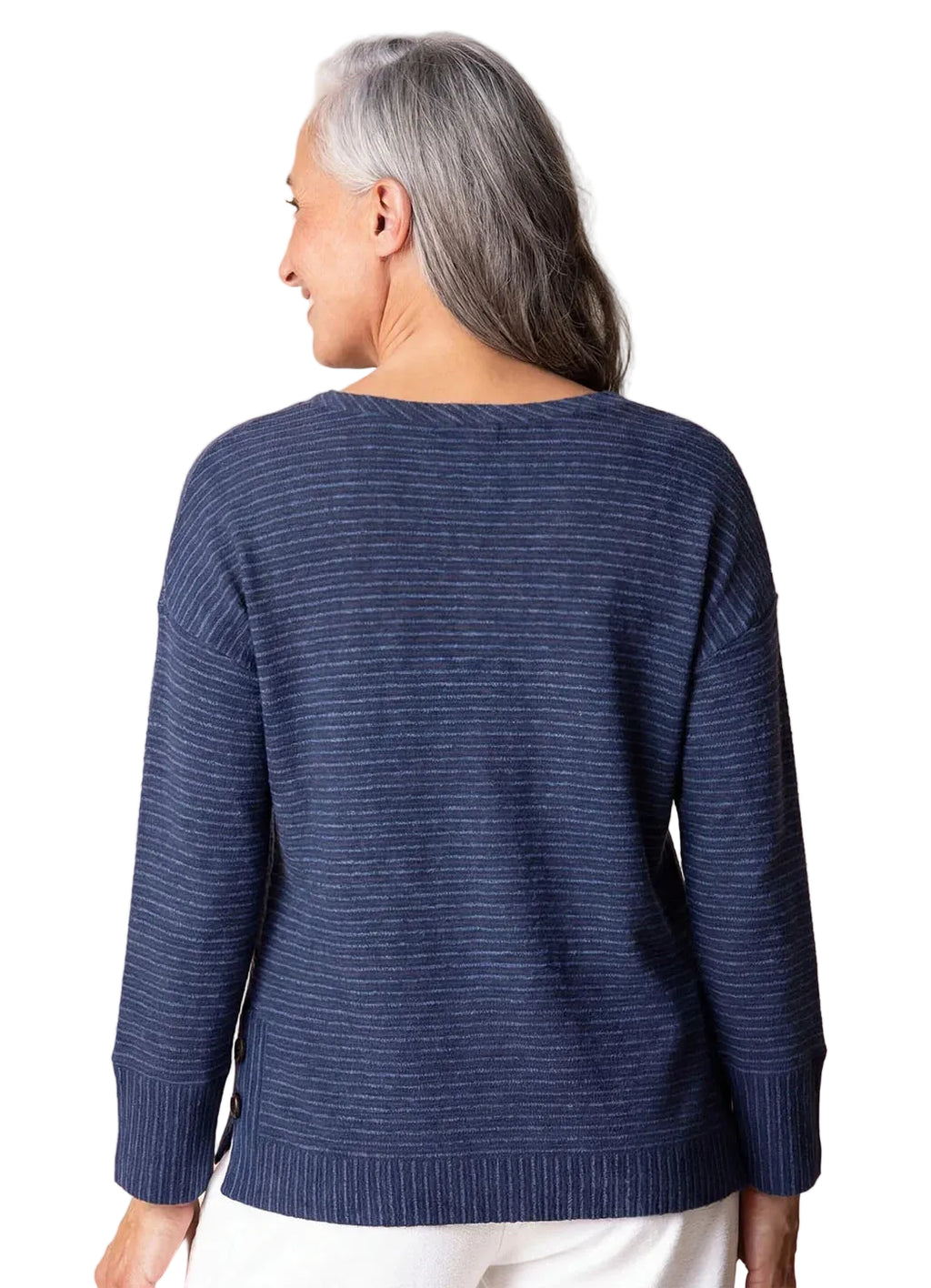 A smiling woman with gray hair wearing a navy blue long-sleeve knit sweater with thin vertical stripes and white pants, standing against a solid beige background.