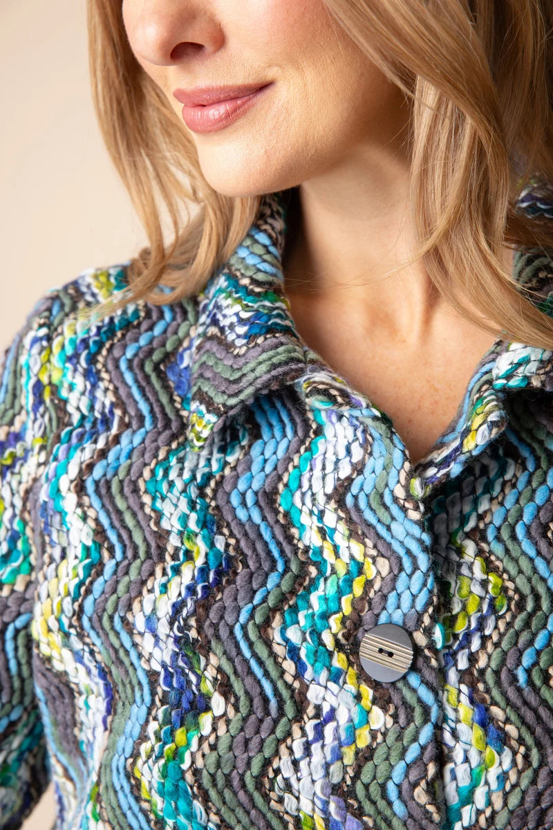 A woman wearing a long-sleeve button-front jacket with a vibrant multicolor zigzag pattern in blue, green, yellow, and gray, paired with dark blue jeans, set against a plain light gray background.
