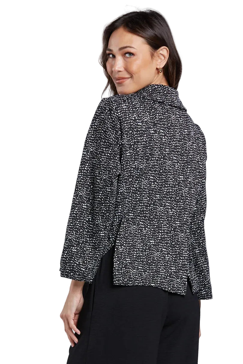 A woman modeling a black and white speckled long-sleeve button-up blouse with a pointed collar, paired with black trousers, against a light gray studio background.